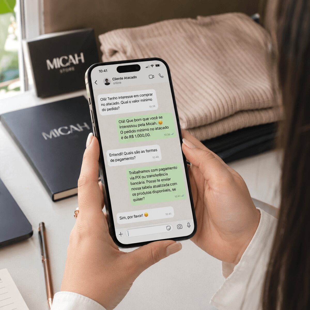 Mãos de uma mulher segurando o celular conversando pelo WhatsApp ao lado de caixas da Micah